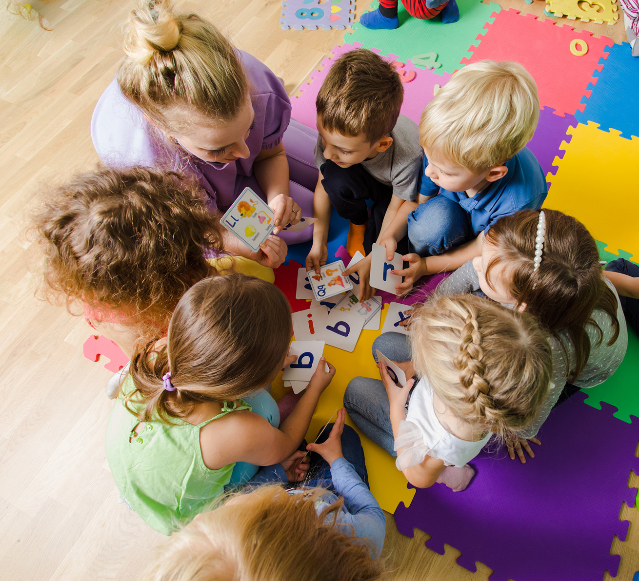 Grupa dzieci z przedszkola siedząca blisko siebie na podłodze razem z nauczycielem prowadzącym pracę w grupie; dzieci uczą się współpracy podczas rozwiązywania zadań